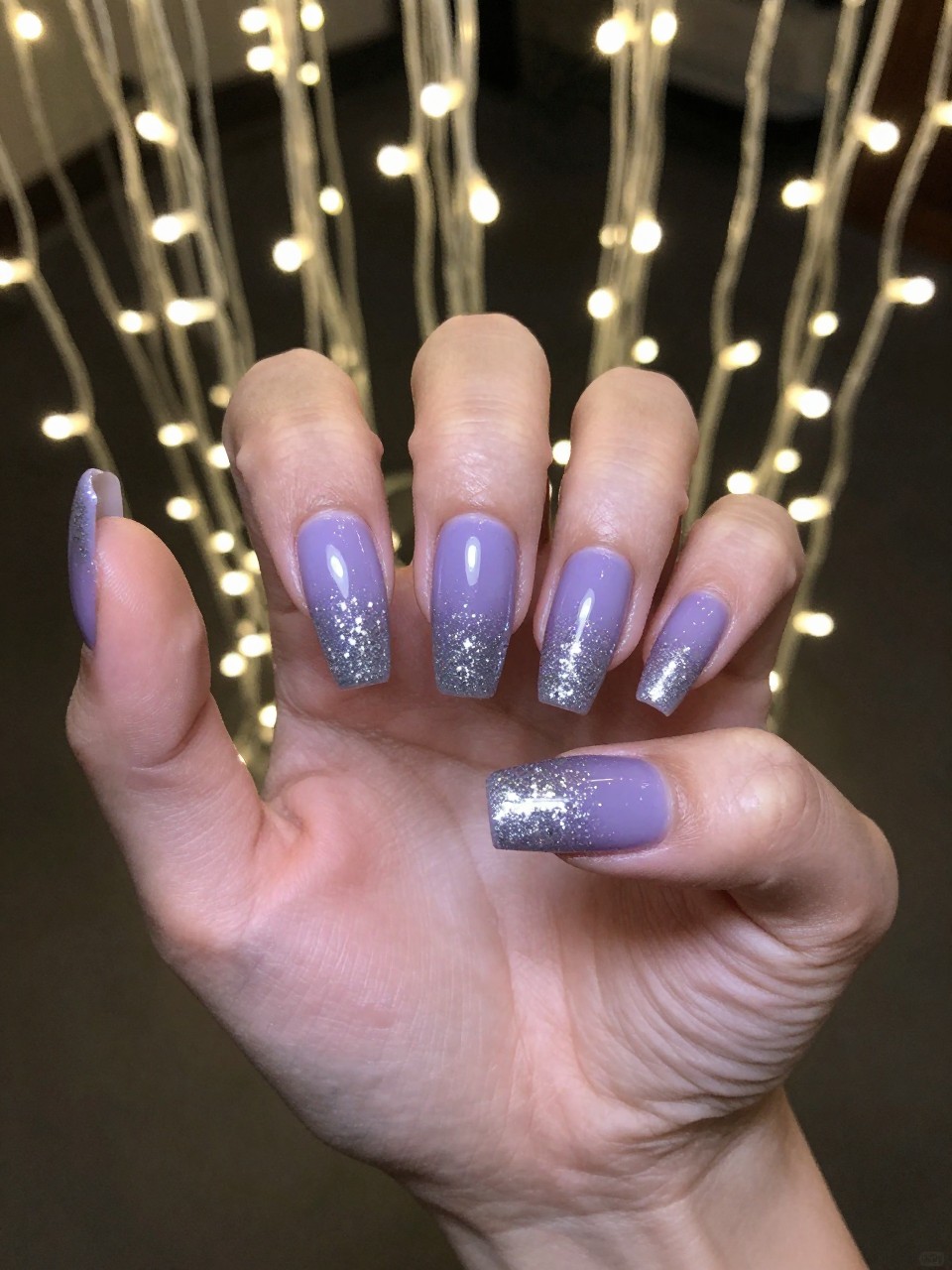 Bird's-eye view of hands under twinkling string lights. The coffin-shaped nails have a gradient from a solid lavender base to dense, silver glitter tips. The dark, evening-inspired background makes the glitter look like a cascade of stars.