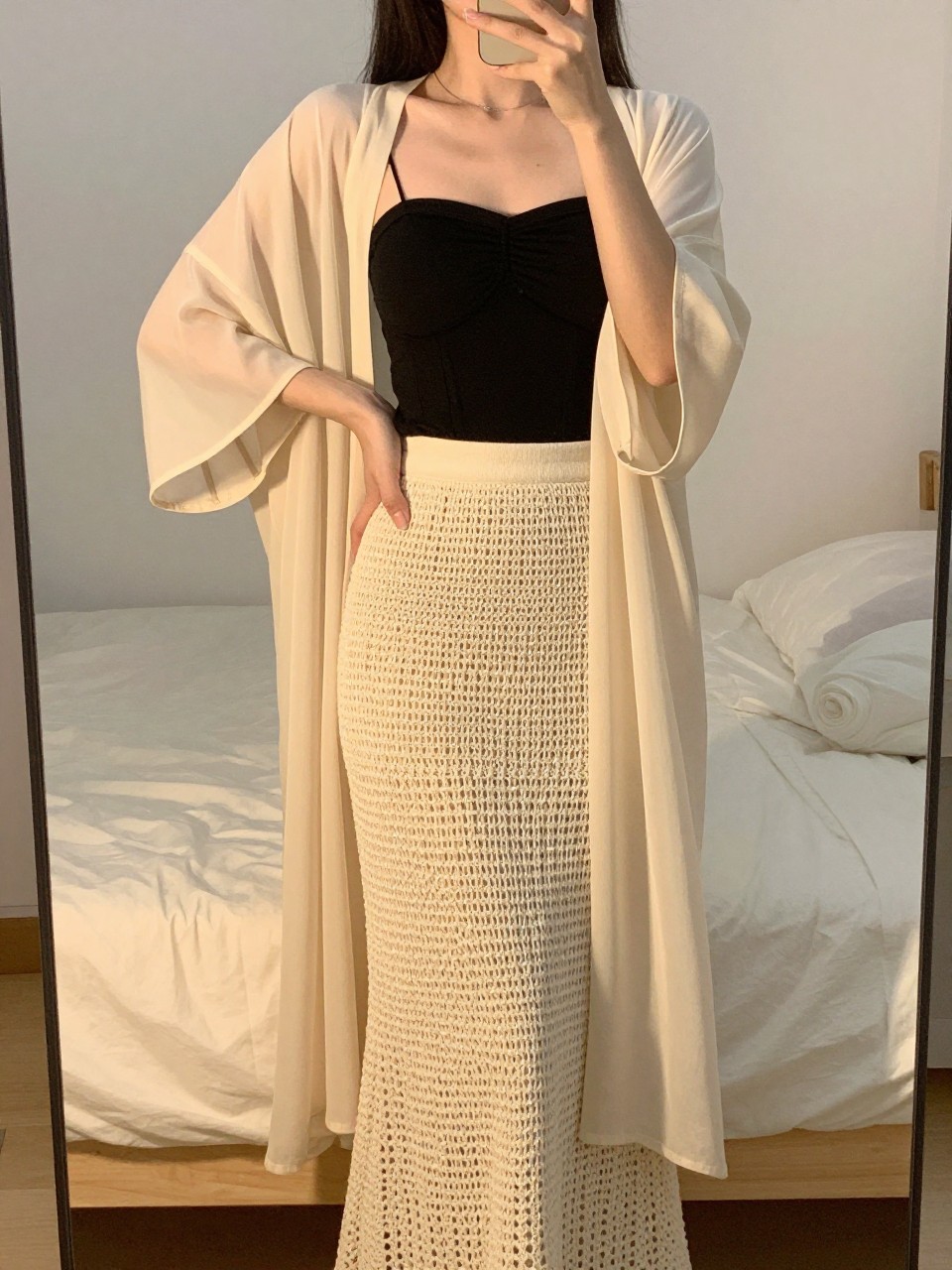 iPhone mirror selfie of a woman wearing a black bandeau top with a natural crochet maxi skirt and a lightweight sheer cream kimono over it, one hand on her hip, cropped at NECK so face doesn't show, in a feminine-themed bedroom, evening golden hour, iPhone photo quality.