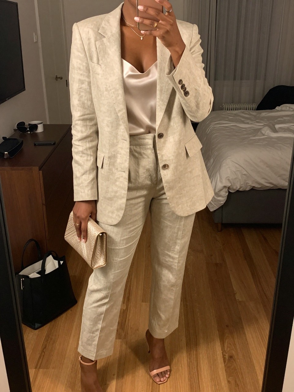 mirror selfie of a black woman wearing a light beige linen blend blazer with matching tailored trousers and an ivory satin cami, nude strappy heels, and a champagne small clutch, holding a phone in front of a mirror, cropped at NECK so face doesn't show, with a natural casual selfie pose, in a city apartment, warm moody lighting, including small lived-in details like a tote bag on the floor, sunglasses on a dresser, or soft bedding in the background, latest iPhone photo quality.