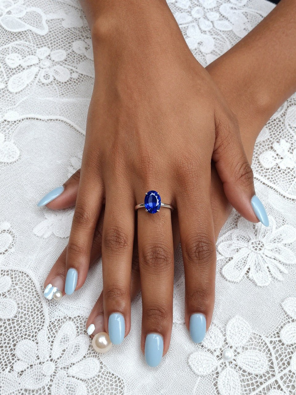 Overhead shot of black feminine hands with a delicate blue sapphire ring, resting on a white lace cloth. The nails are a short squoval shape in a milky light blue. A single pearl sits at the cuticle of each ring finger. The lighting is bright and cheerful.