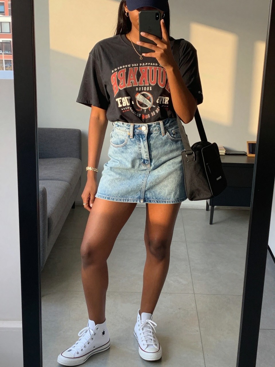 mirror selfie of a black woman wearing a black vintage band tee tucked into a light-wash denim mini skirt with white Converse sneakers and a baseball cap, holding a black canvas crossbody bag, standing in a casual classic pose, cropped at NECK so face doesn't show, in a city apartment, evening golden hour, iPhone photo quality.