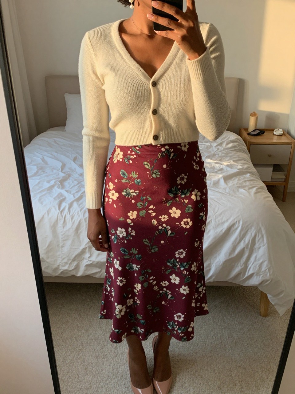 mirror selfie of a black woman wearing a cream cropped cardigan with a maroon floral skirt and nude heels, one hand gently touching her pearl earring, cropped at NECK so face doesn't show, in a feminine-themed bedroom, evening golden hour, iPhone photo quality.