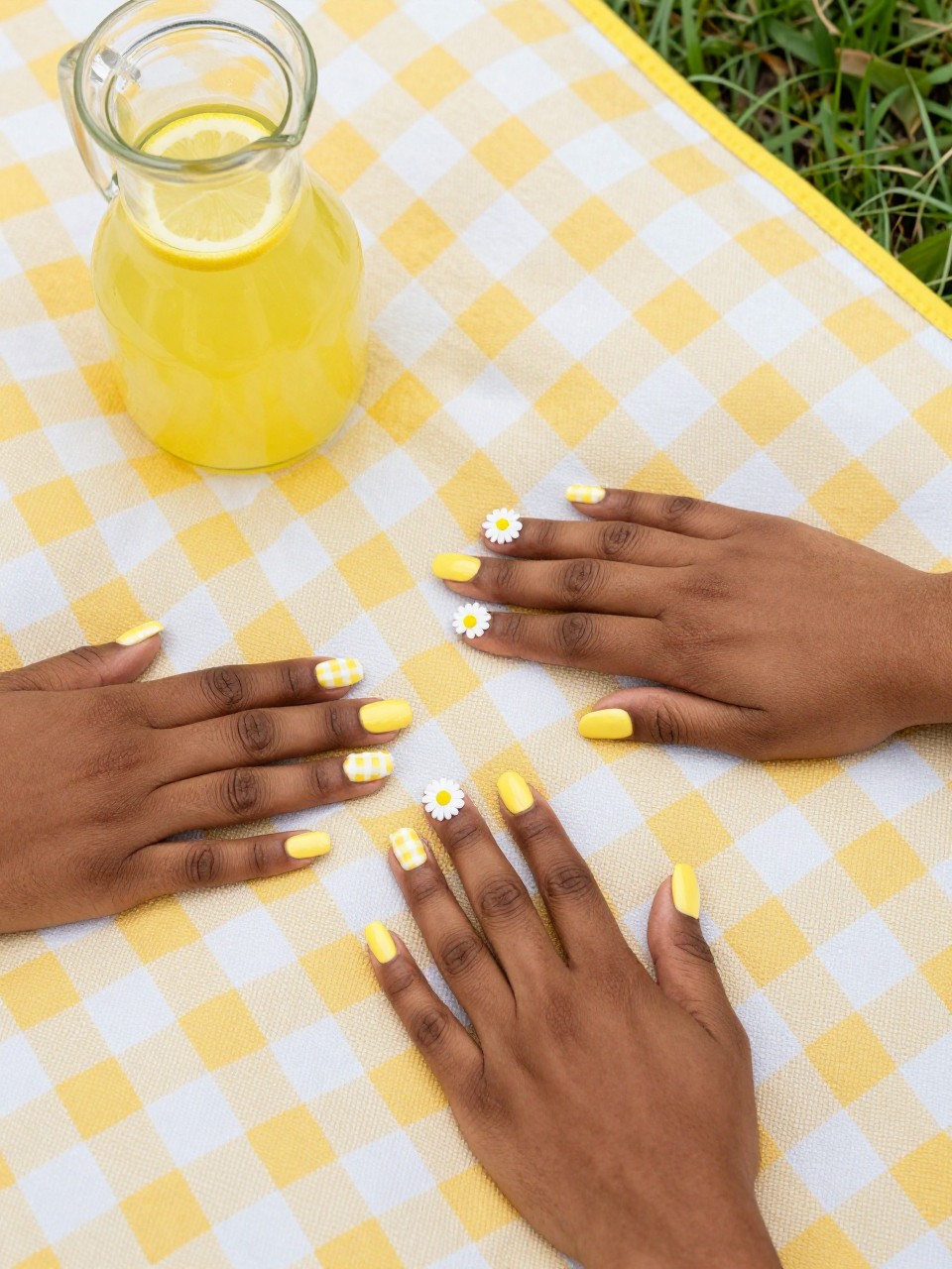 Top-down view of black feminine hands with short nails on a yellow and white gingham picnic blanket. Some nails are yellow and white checkered. The accent nails are solid yellow with one bold white daisy. A lemonade pitcher sits nearby in the grass.