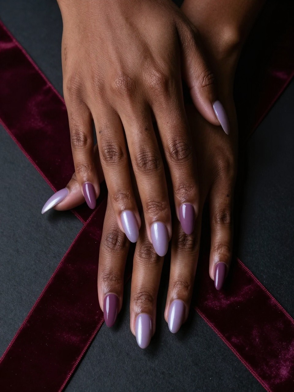 Top-down view of black feminine hands resting on a burgundy velvet ribbon. The almond nails showcase a gradient from a deep mauve at the cuticles to a pale, silvery lilac at the tips. Moody, directional lighting highlights the depth of the colors.