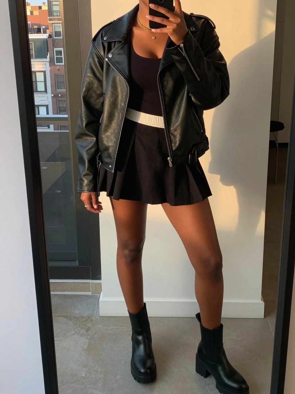 mirror selfie of a black woman wearing a black leather jacket over a black tank top with a black tennis skirt and chunky ankle boots, one hand in her jacket pocket, cropped at NECK so face doesn't show, in a city apartment, evening golden hour, iPhone photo quality.