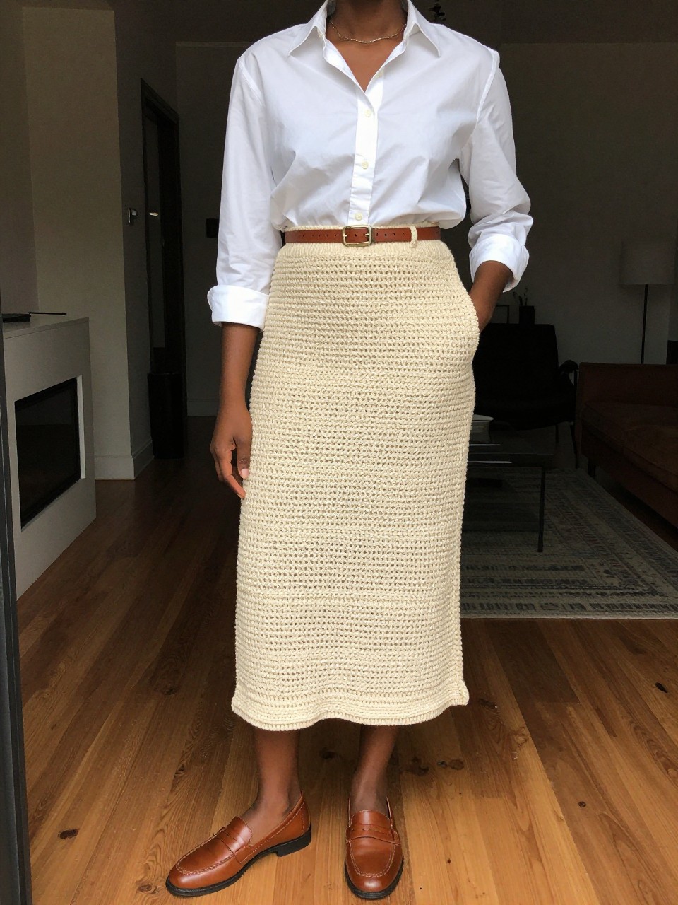 photo of a black woman wearing a white button-down shirt tucked into a natural crochet midi skirt with brown leather loafers and a matching belt, one hand in her pocket, cropped at NECK so face doesn't show, in a city apartment, warm moody lighting, iPhone photo quality.