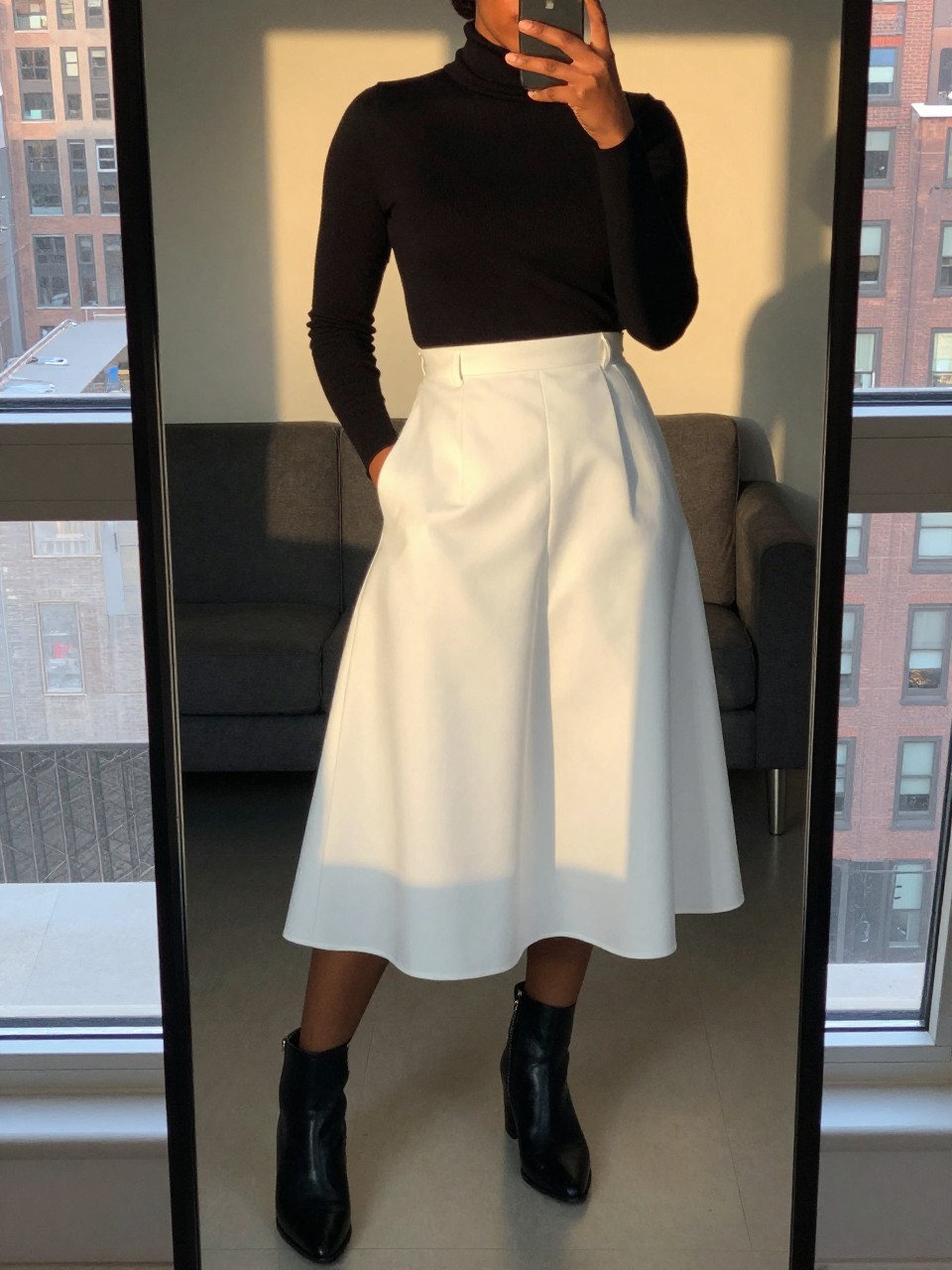 mirror selfie of a black woman wearing a black turtleneck tucked into a white A-line skirt with black ankle boots, her hair in a low bun, one hand in her pocket, cropped at NECK so face doesn't show, in a city apartment, evening golden hour, iPhone photo quality.