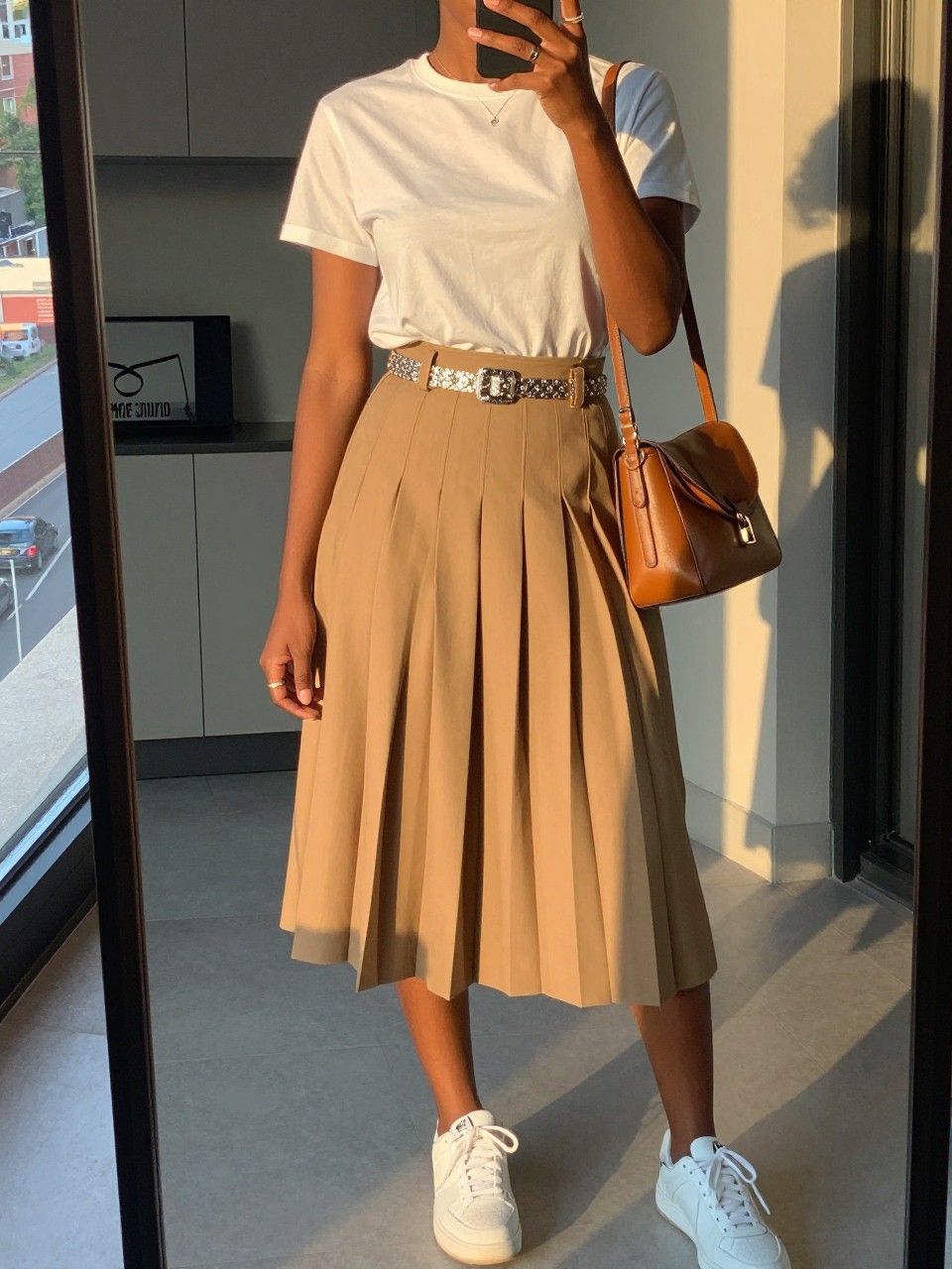 iPhone mirror selfie of a black woman wearing a white crewneck t-shirt tucked into a camel pleated midi skirt with a silver statement belt at her waist and white sneakers, holding a tan leather crossbody bag, standing in a casual elegant pose, cropped at NECK so face doesn't show, in a city apartment, evening golden hour, iPhone photo quality.