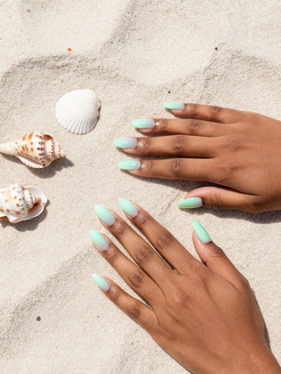 Overhead shot of black feminine hands placed near seashells on a light sand-colored towel. The squoval nails feature a vertical ombré from a pale mint green to a translucent seafoam. The bright, natural light evokes a sunny day at the shore.