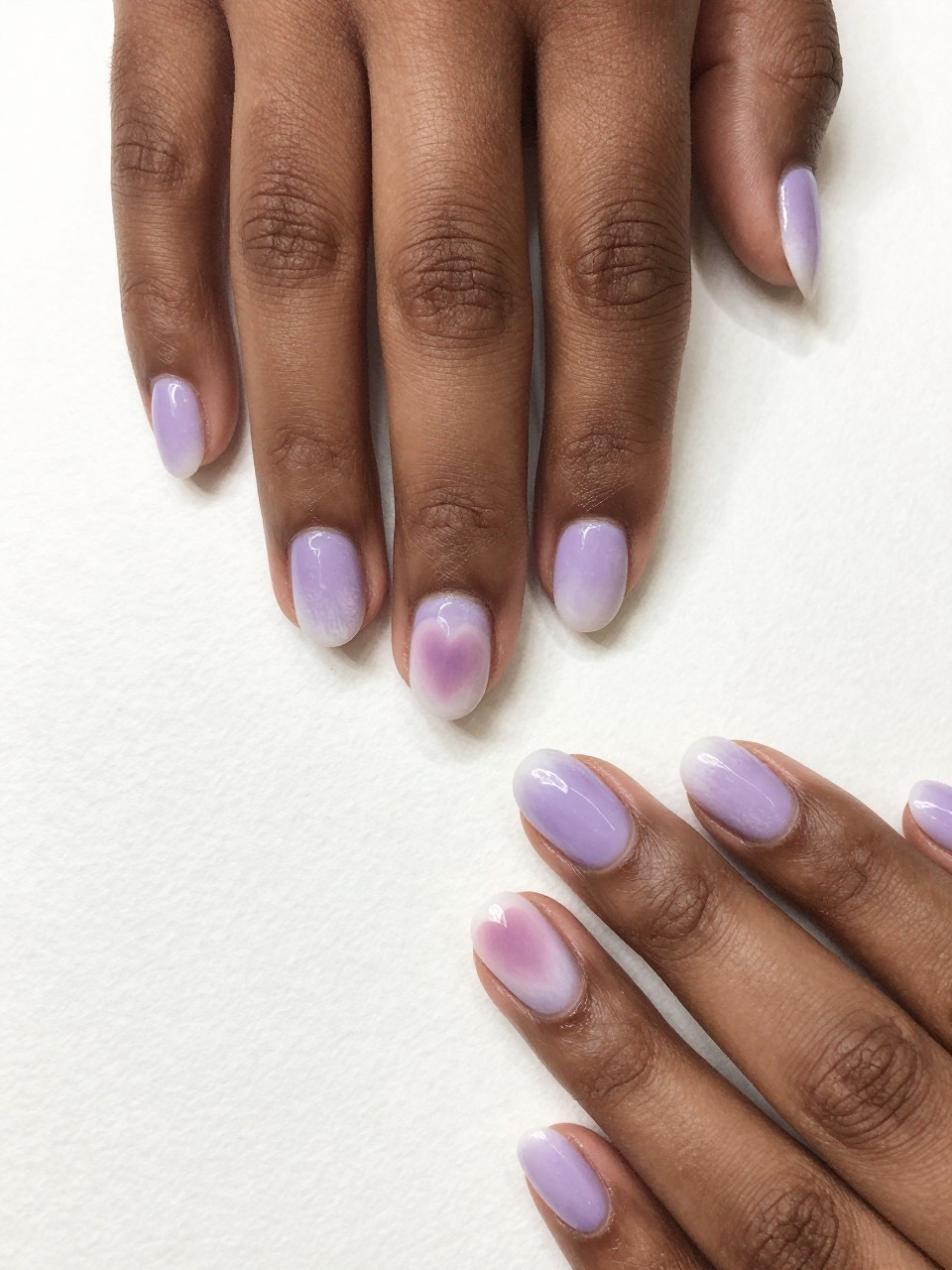Top-down view of black feminine hands with oval nails on watercolor paper. The nails have a soft lavender and white wash. A blurred, heart-shaped blend of color sits in the center of each nail. The style is painterly, soft-focus, and romantic.