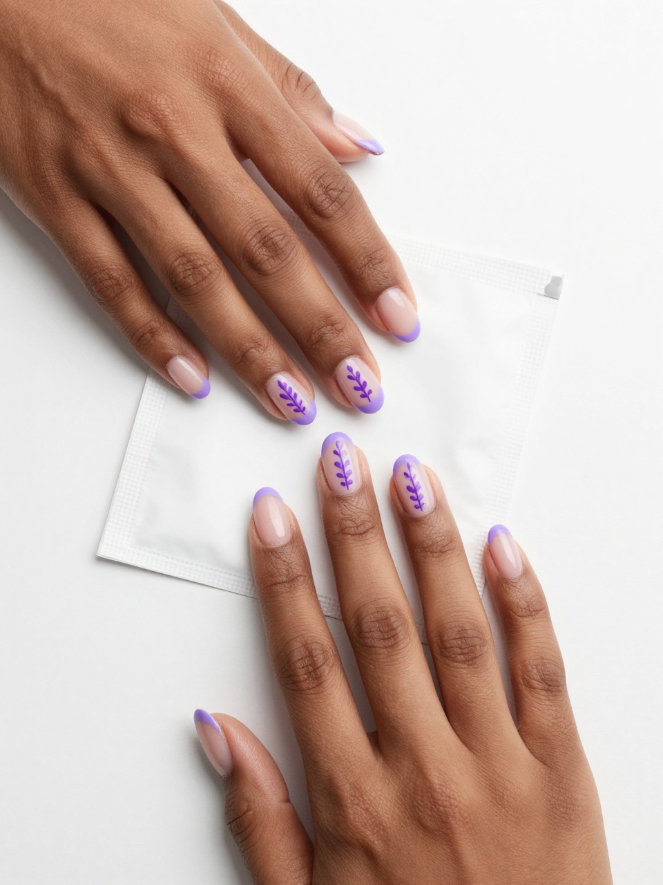 Overhead shot of black feminine hands with oval nails resting on a sachet. The nails have lavender French tips. on the nails, there is a single vertical lavender sprig that is painted on the sheer base of each nail, extending from the tip. The mood is calm and unified.