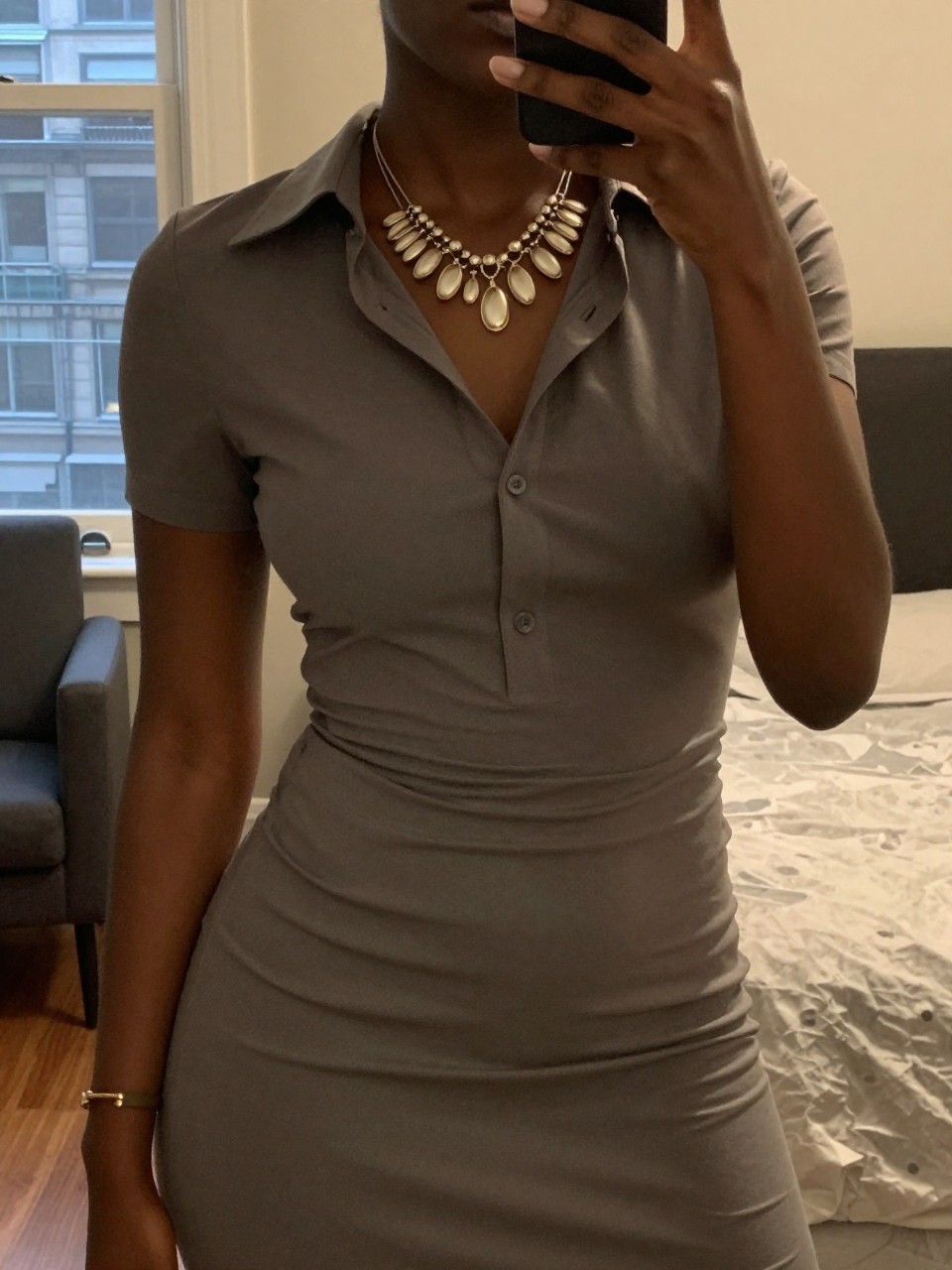 mirror selfie of a black woman in a shirt dress with a statement necklace, touching the necklace, cropped at NECK so face doesn't show, in a city apartment, warm moody lighting, iPhone photo quality.
