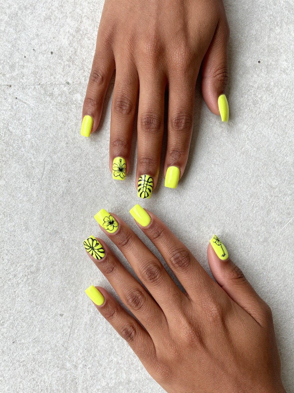 Overhead shot of black feminine hands with square nails against a concrete wall. The nails are matte neon yellow, and it features a bold, black outlined monstera leaves or hibiscus flowers are painted on each nail. The vibe is urban and artistic.