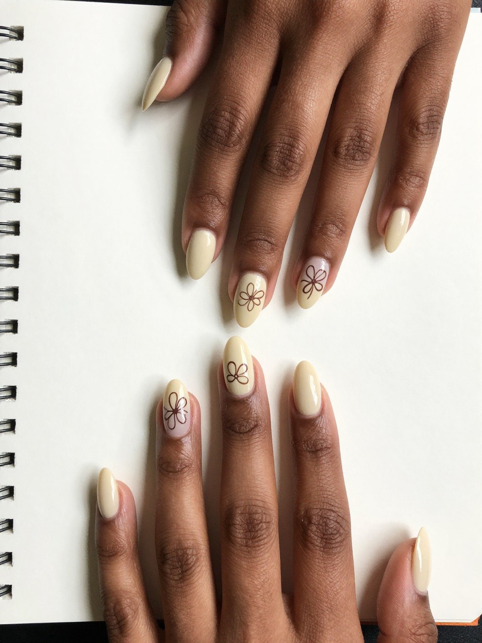 Top-down view of black feminine hands with almond nails resting on a page of a sketchbook. The nails are a matte cream color. Each nail features a simple, hand-drawn brown outline of a flower, with intentionally imperfect lines. The mood is artistic, warm, and casual.