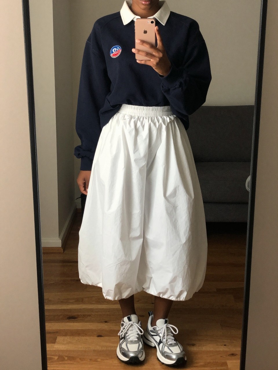 iPhone mirror selfie of a black woman in a navy rugby shirt and a white bubble skirt with dad sneakers, casually holding her phone at chest level, cropped at NECK so face doesn't show, in a city apartment, warm moody lighting, iPhone photo quality.