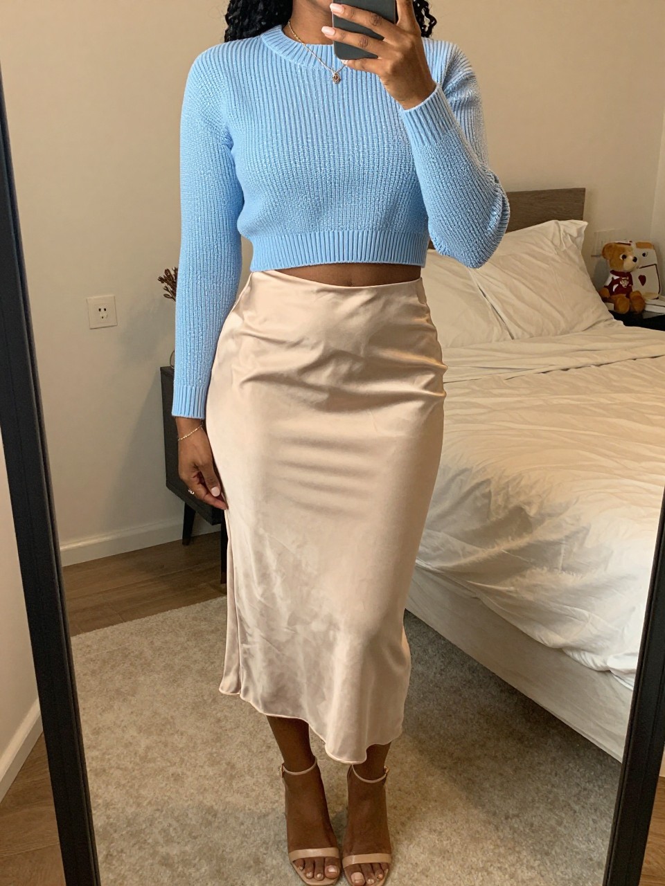 iPhone mirror selfie of a black woman wearing a baby blue cropped ribbed sweater with a high-waisted champagne satin skirt and nude block-heel sandals, one hand gently touching her necklace, cropped at NECK so face doesn't show, in a feminine-themed bedroom, warm moody lighting, iPhone photo quality.