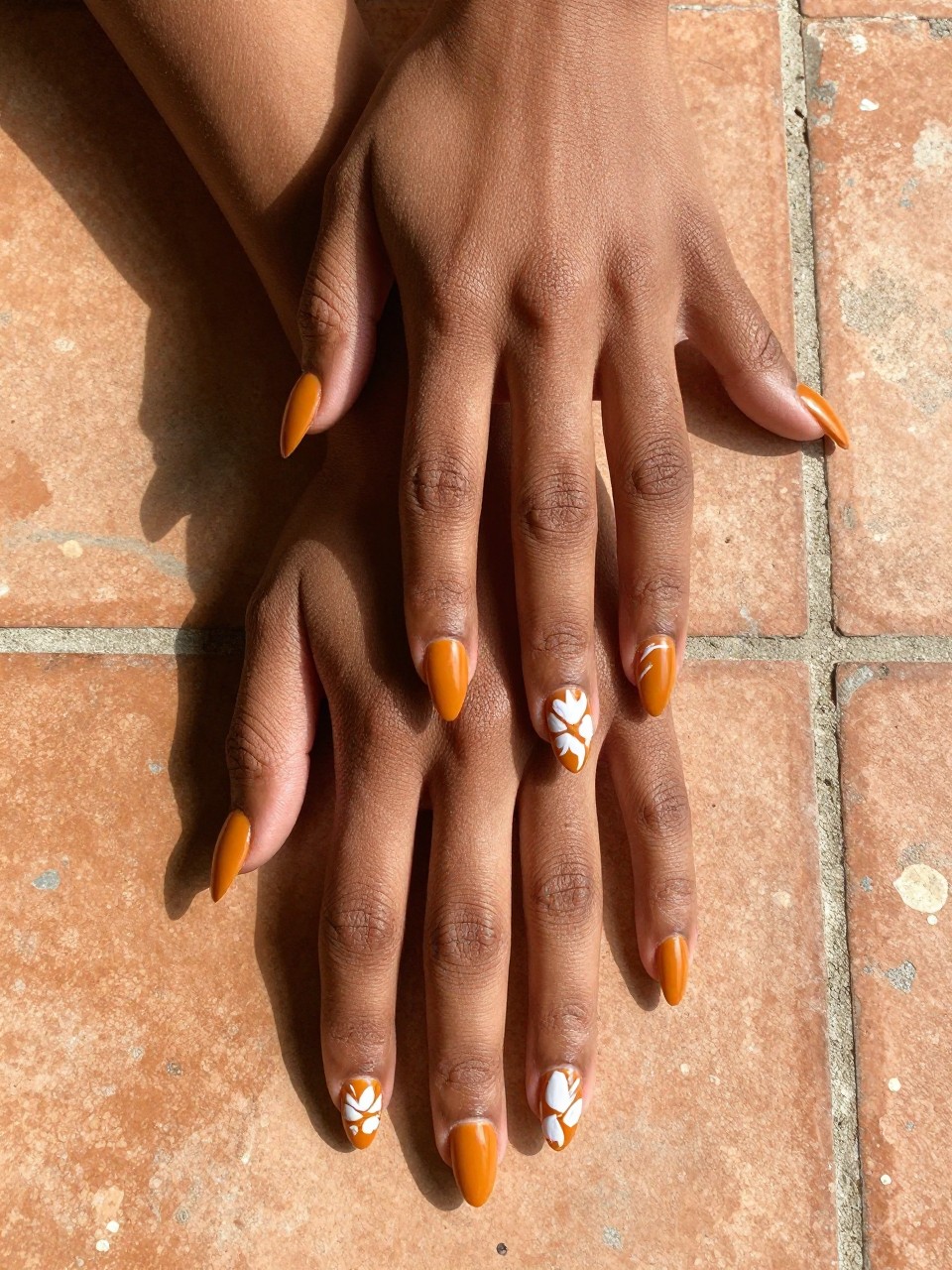 Bird's-eye view of black feminine hands with almond nails resting on a rustic terracotta tile. The nails are a matte marigold orange. Each nail features unique, abstract white floral brushstrokes. The setting feels like an artist’s sunlit studio.