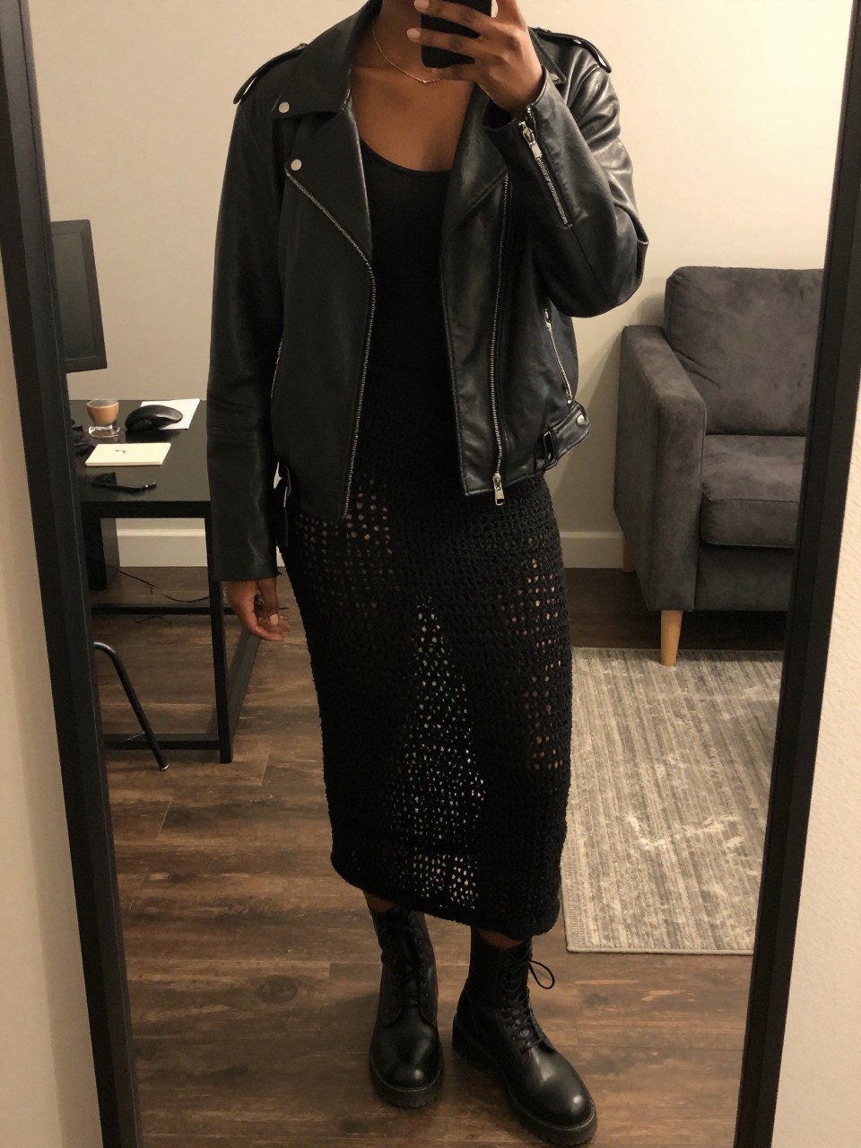 mirror selfie of a black woman wearing a black leather jacket over a black tank top with a black crochet midi skirt and black combat boots, one hand in her jacket pocket, cropped at NECK so face doesn't show, in a city apartment, warm moody lighting, iPhone photo quality.