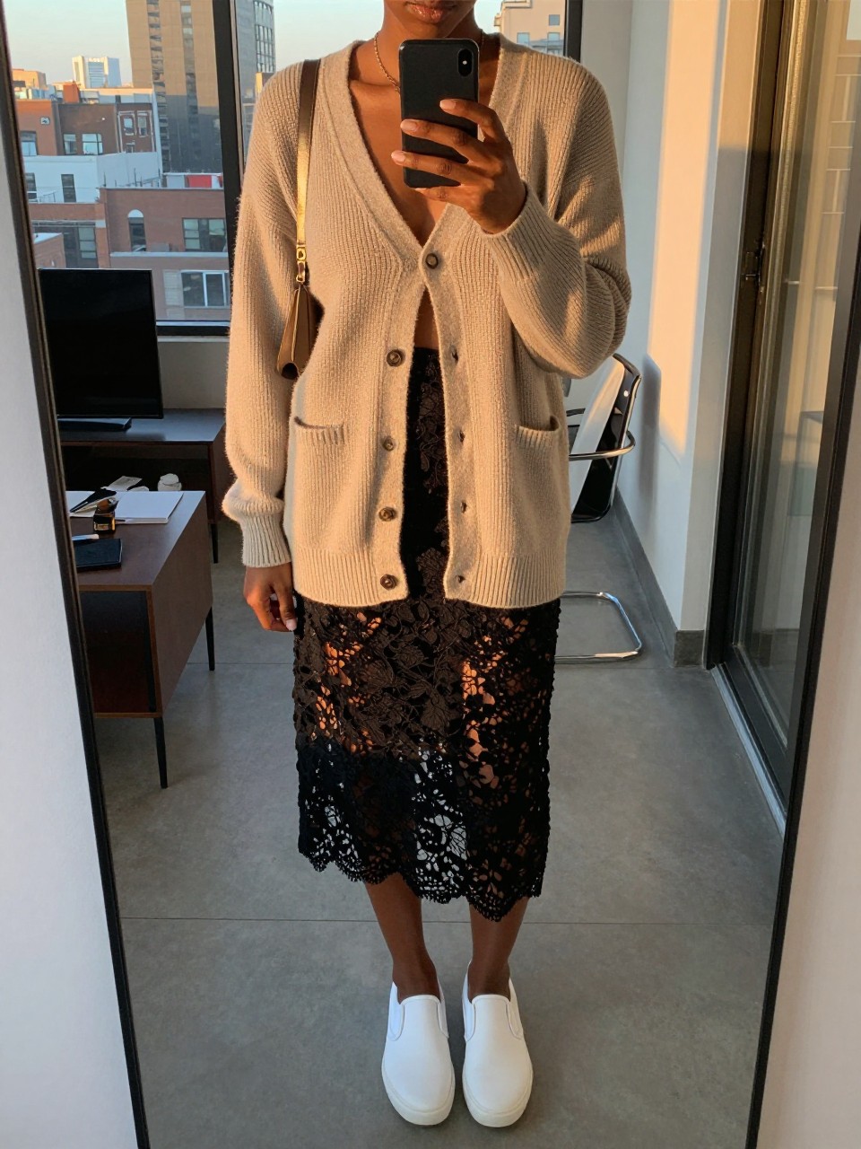 mirror selfie of a black woman in an oatmeal cardigan and black lace skirt with white slip-on sneakers, phone in one hand, cropped at NECK so face doesn't show, in a city apartment, evening golden hour, iPhone photo quality.