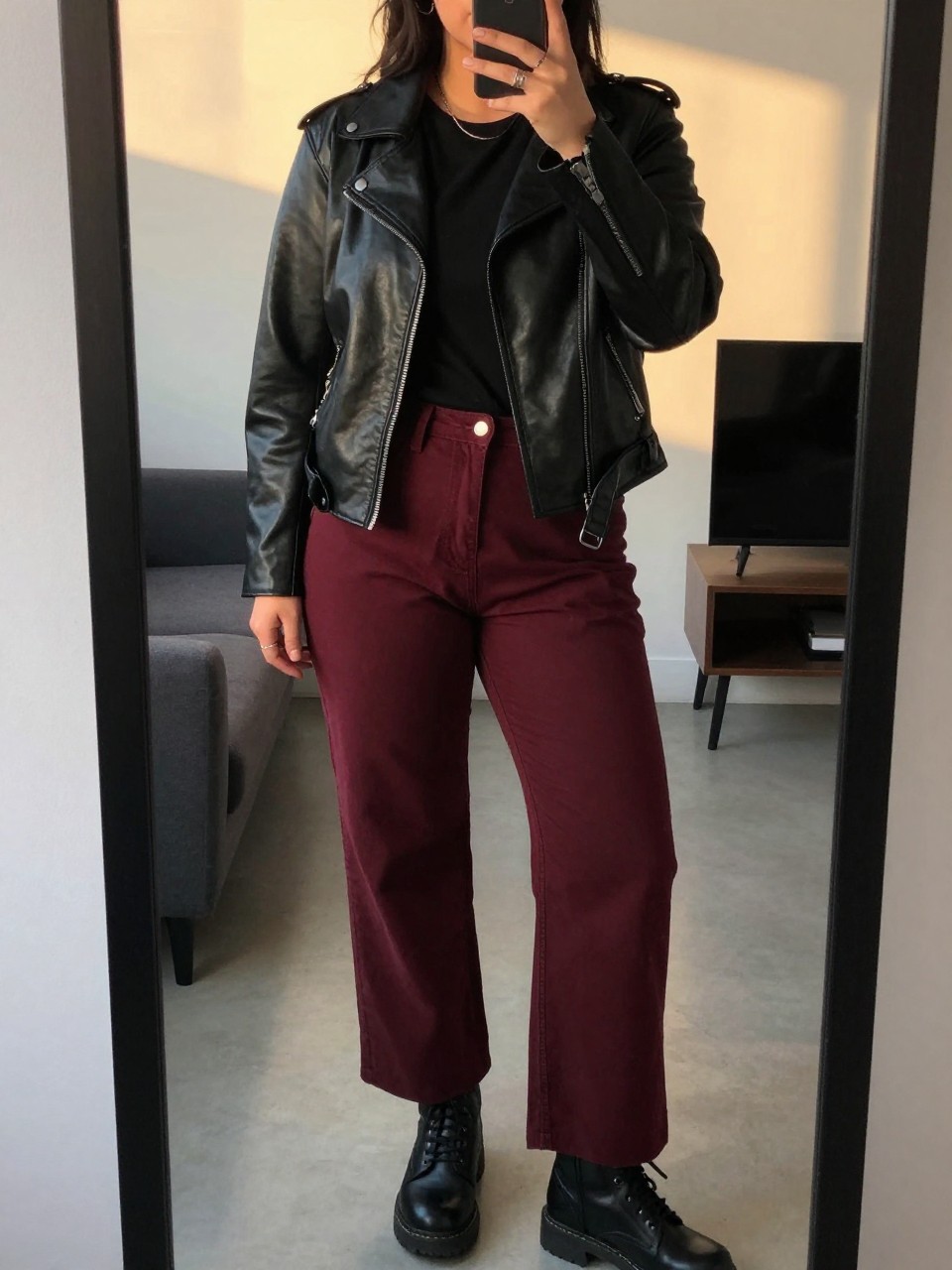 mirror selfie of a curvy woman in a black leather jacket over a tee with burgundy wide leg pants and combat boots, one hand in jacket pocket, cropped at NECK so face doesn't show, in a city apartment, evening golden hour, iPhone photo quality.