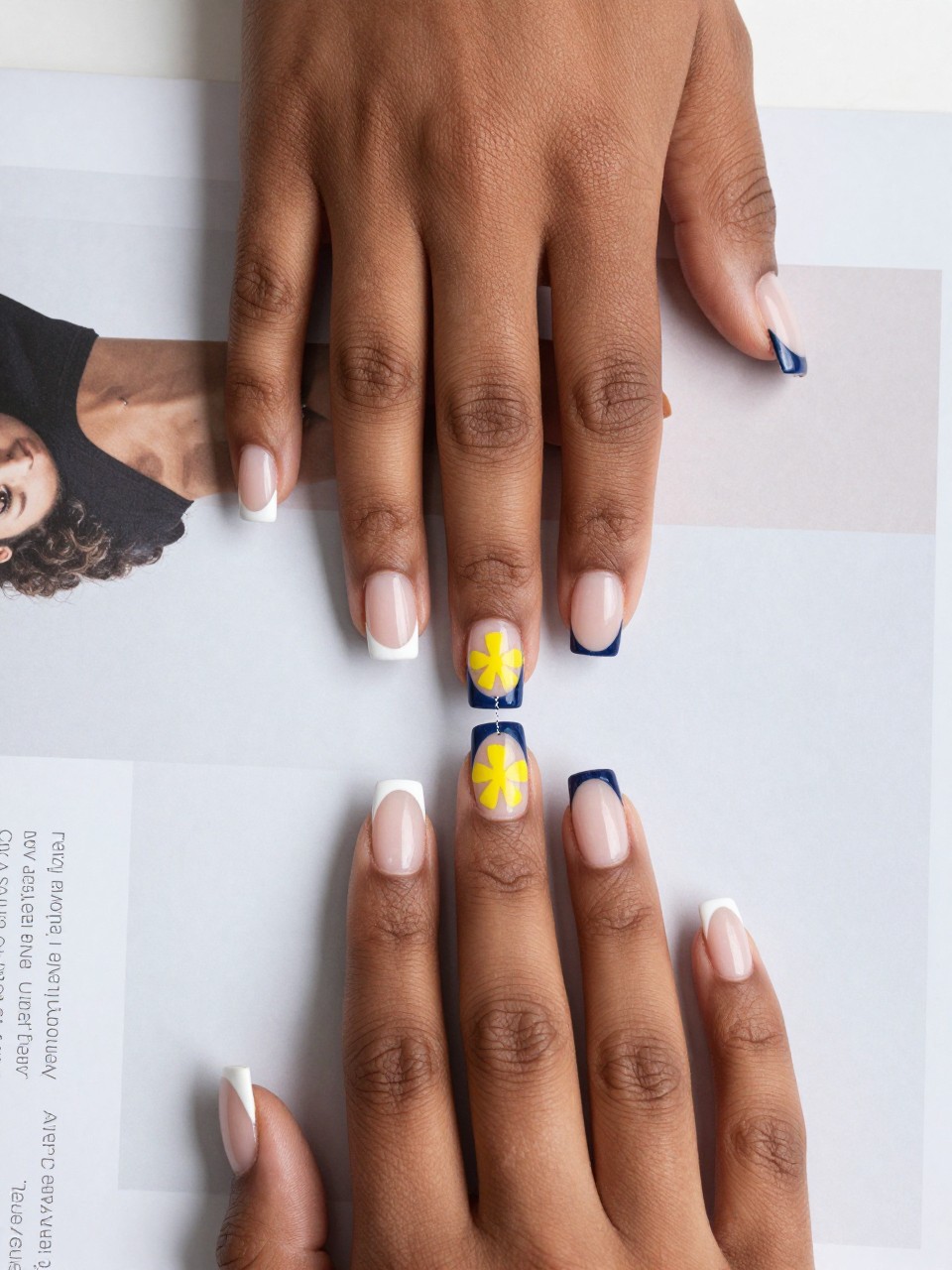 Overhead view of black feminine hands with square nails on a modern design magazine. The French tips are split vertically into white and navy. A small, geometric yellow flower is painted at the seam on the nail bed. The look is crisp and contemporary.