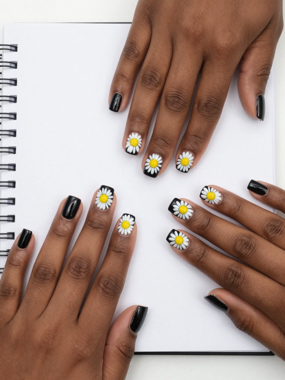 Overhead view of black feminine hands with short, squared nails on a white notebook. The nails are a glossy, deep black. Bold white daisies with bright yellow centers are painted on each nail. The contrast is stark, clean, and eye-catching.