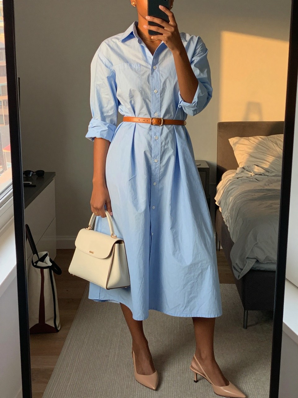 mirror selfie of a black woman wearing a light blue cotton poplin midi shirt dress with a tan leather thin waist belt, nude leather slingback heels, a cream structured handbag, and simple gold stud earrings, holding a phone in front of a mirror, cropped at NECK so face doesn't show, with a natural casual selfie pose, in a city apartment, evening golden hour, including small lived-in details like a tote bag on the floor, sunglasses on a dresser, or soft bedding in the background, latest iPhone photo quality.