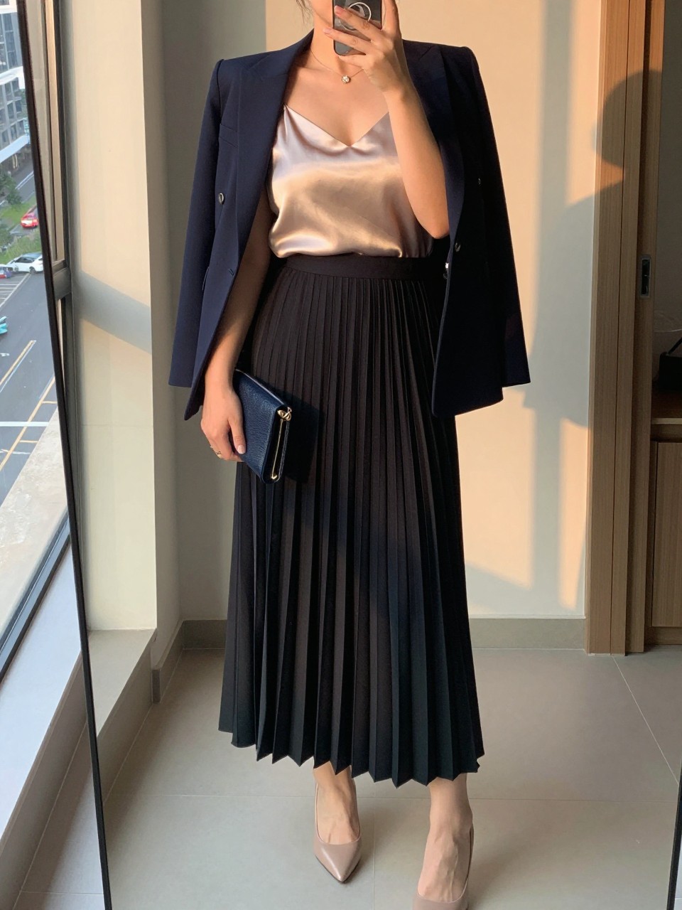 iPhone mirror selfie of a curvy woman wearing a champagne silk camisole tucked into a black pleated maxi skirt with a structured navy blazer worn open and nude pointed heels, holding a small navy clutch, standing in a polished professional pose, cropped at NECK so face doesn't show, in a city apartment, evening golden hour, iPhone photo quality.