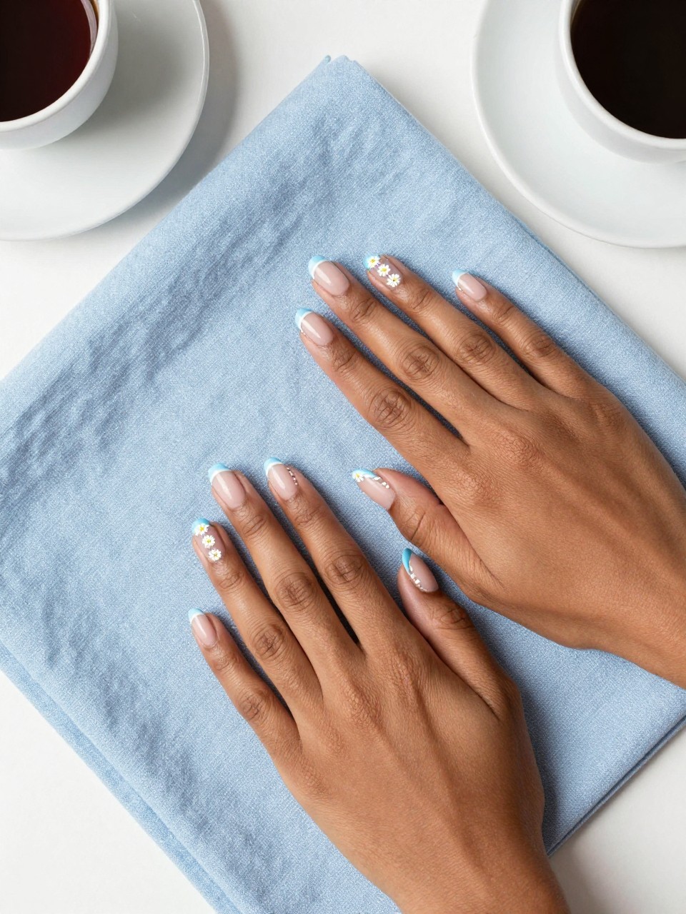 Bird's-eye view of black feminine hands with short, rounded nails resting on a light blue linen cloth. The nails have baby blue French tips. on the nails, there are chains of micro white daisies traces the line of the blue tip. The setting is a bright, sunlit breakfast table.