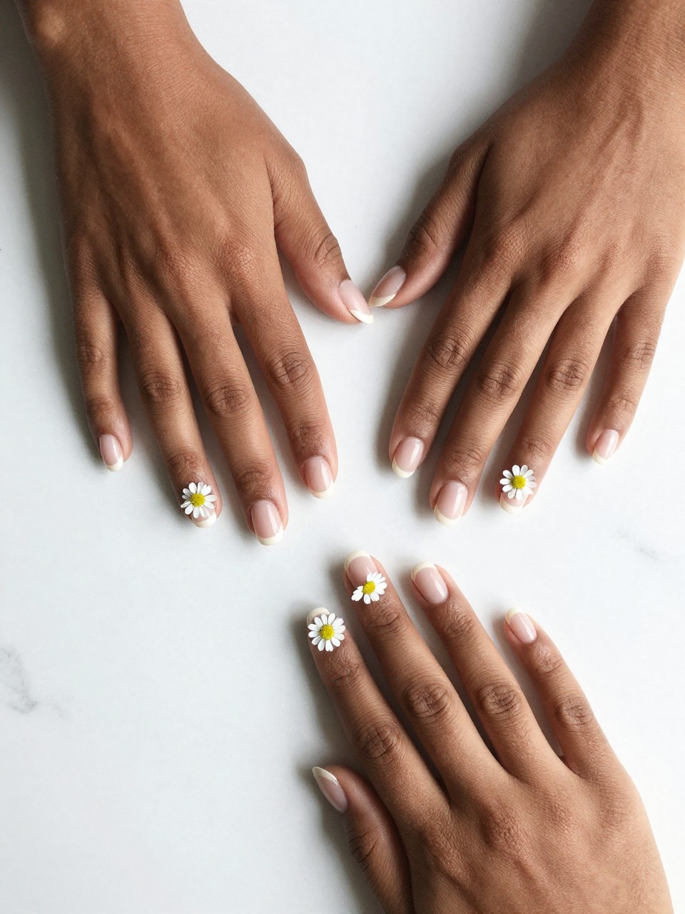 Overhead shot of black feminine hands with neat, short nails on a white marble counter. The nails have a sheer nude base. A single line of connected tiny white daisies wraps around each nail like a delicate chain. The composition is crisp and minimalist.