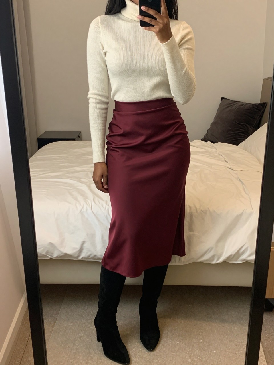 iPhone mirror selfie of a black woman wearing a cream turtleneck tucked into a burgundy satin midi skirt with black tights and black suede knee-high boots, one hand adjusting her sleeve, cropped at NECK so face doesn't show, in a feminine-themed bedroom, warm moody lighting, iPhone photo quality.