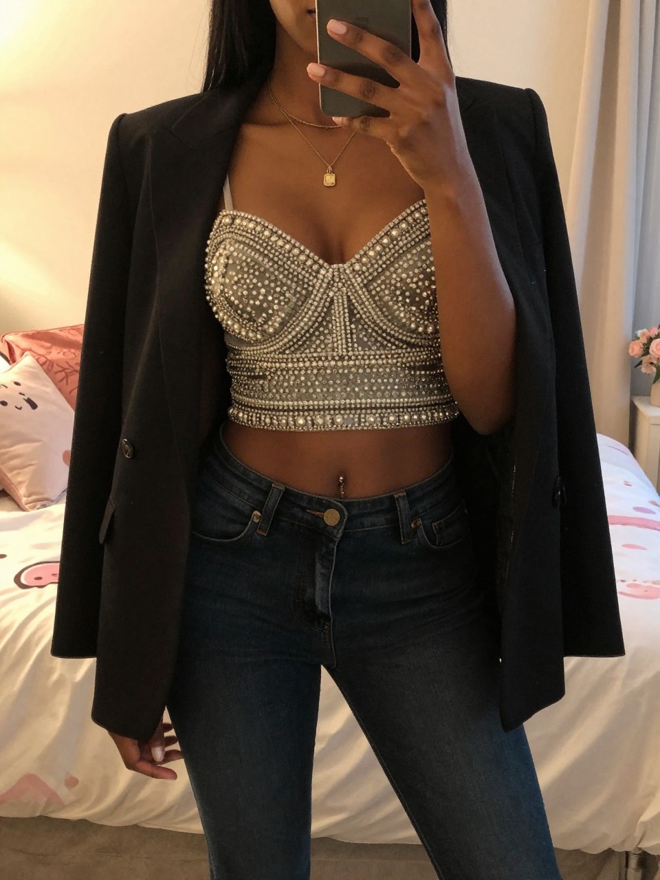 mirror selfie of a black woman in an embellished top and dark jeans, blazer on shoulders, cropped at NECK so face doesn't show, in a feminine-themed bedroom, evening golden hour, iPhone photo quality.