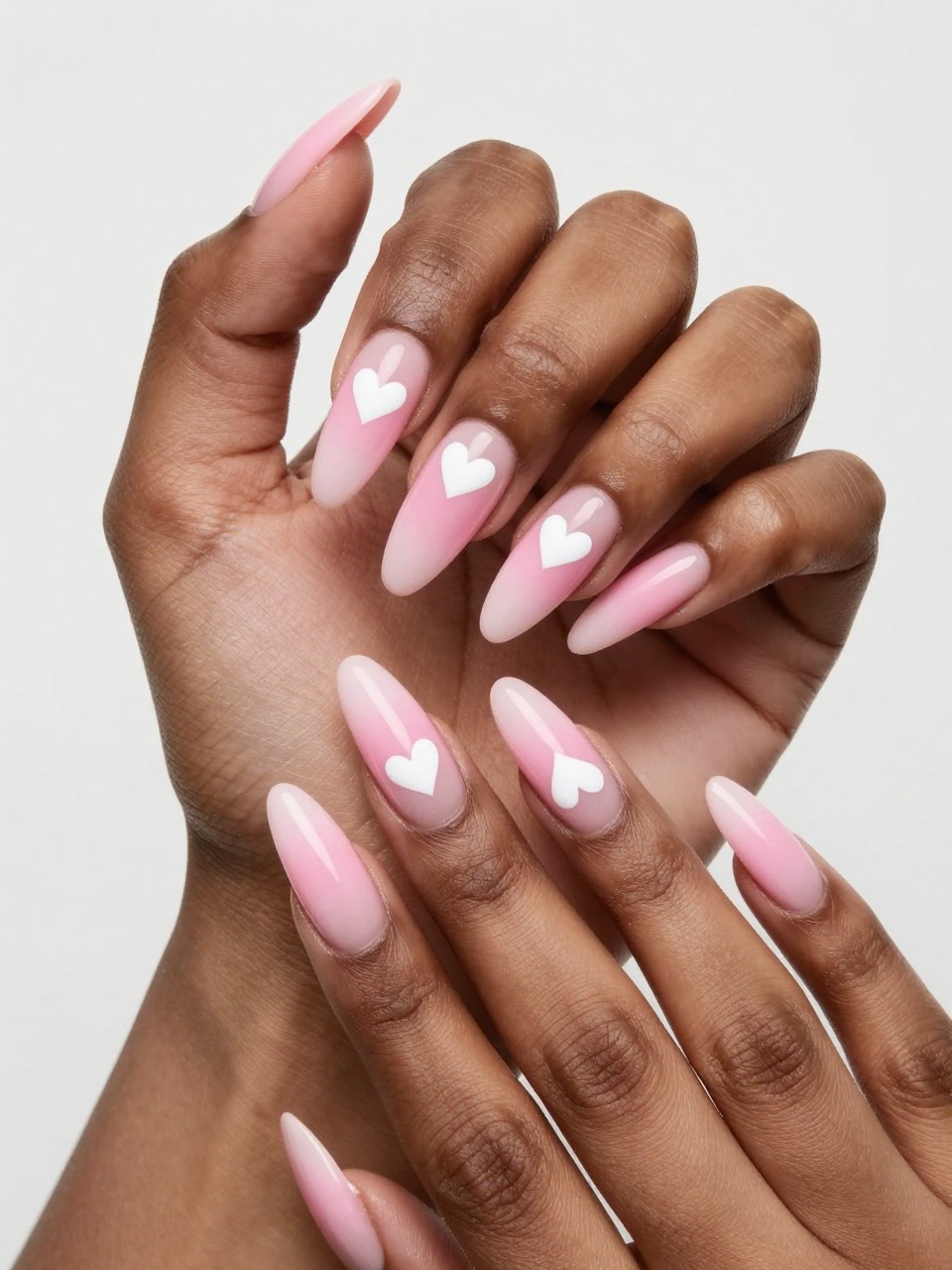 Angled view of black feminine hands with long nails. The nails show a smooth pink ombré fade. Two or three small white hearts are painted on each nail, seemingly suspended in the gradient. The effect is ethereal and romantic.
