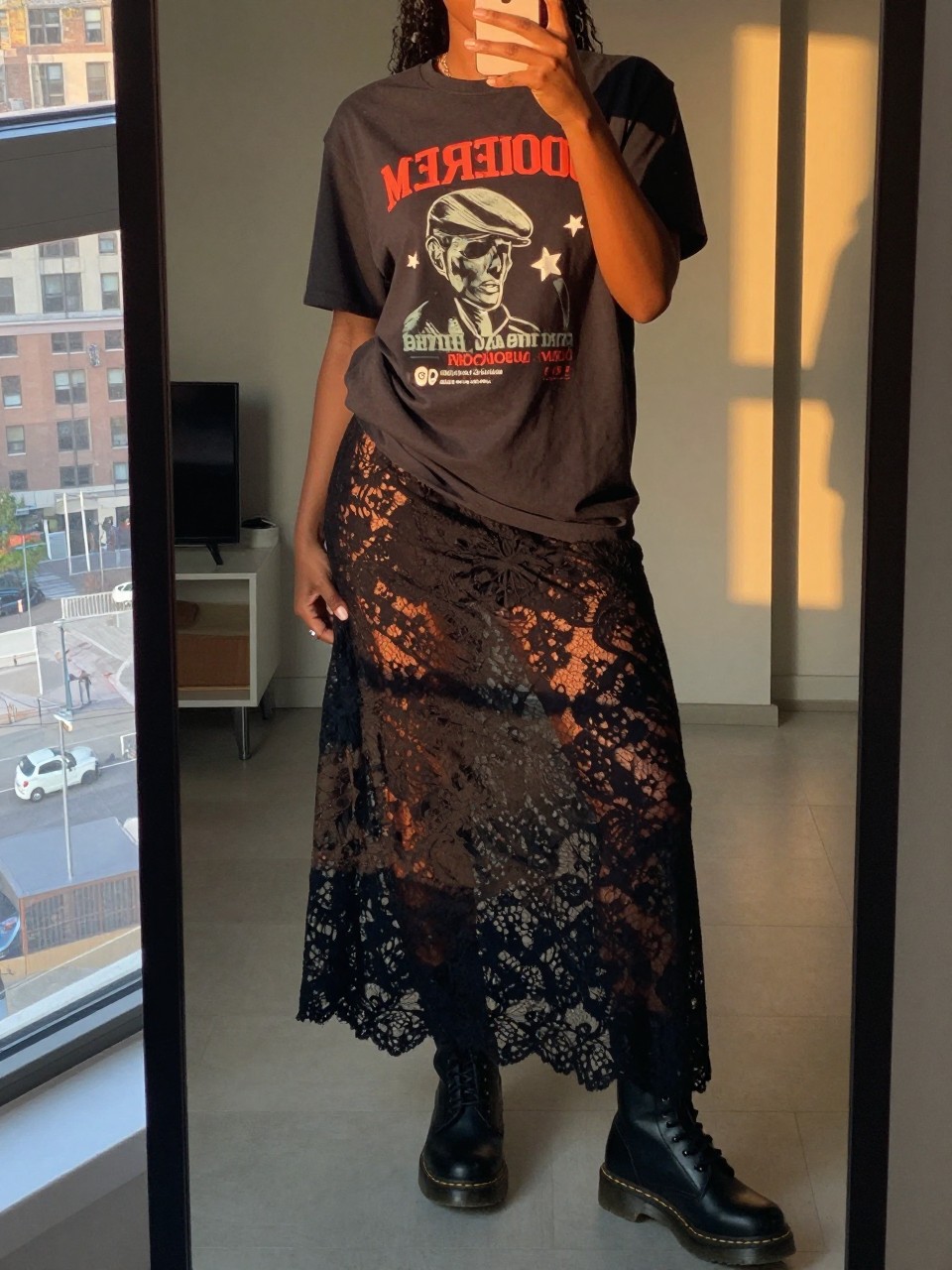 mirror selfie of a black woman in a band tee and black lace maxi skirt with Doc Martens boots, one hand in her pocket, cropped at NECK so face doesn't show, in a city apartment, evening golden hour, iPhone photo quality.