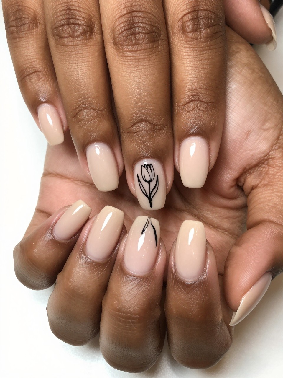 Close-up, top-down view of black feminine hands under bright light. The nail bed is matte beige, the tip is glossy beige. A simple, glossy black line-art tulip is painted on the matte section. The textural difference is sleek and modern.