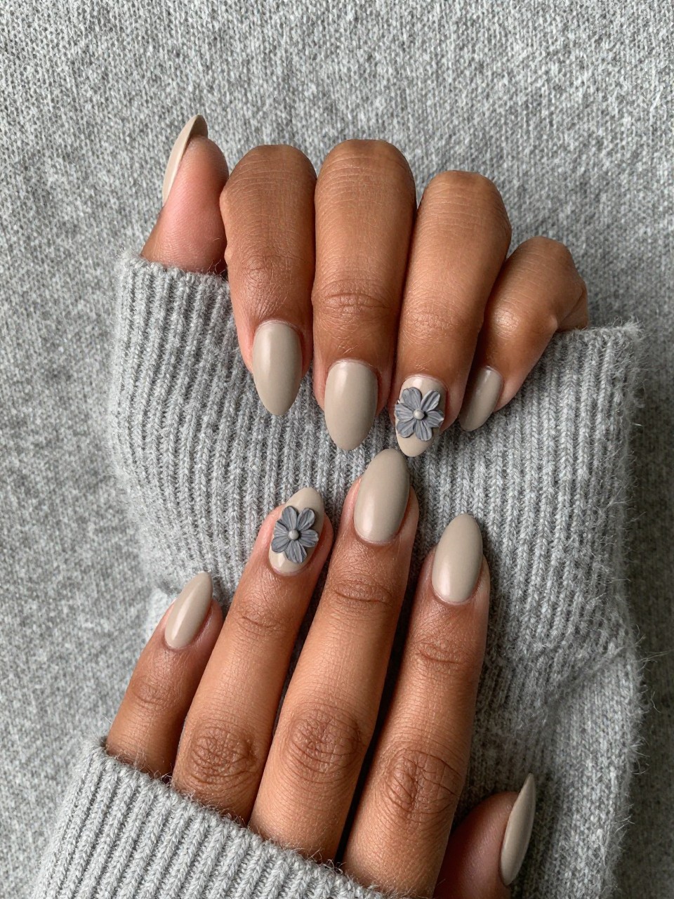 Close-up, top-down view of black feminine hands with almond nails on a grey cashmere sweater. The nails are a matte oatmeal color. Each nail has a single, textured flower painted in a slightly darker matte greige. The look is minimalist, textured, and high-fashion.