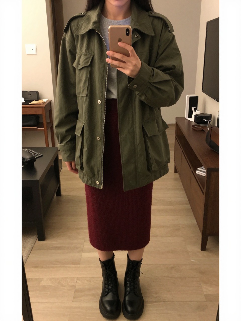 mirror selfie of a woman wearing an olive green military jacket over a gray tee and maroon knit skirt with black combat boots, holding her phone low, cropped at NECK so face doesn't show, in a city apartment, warm moody lighting, iPhone photo quality.