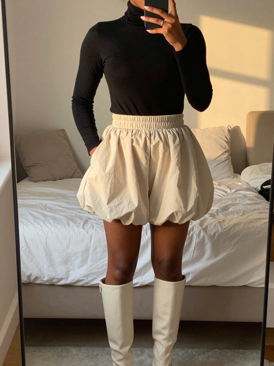 iPhone mirror selfie of a black woman in a black turtleneck and a cream bubble skirt with knee-high boots, standing with one hand in her pocket, cropped at NECK so face doesn't show, in a feminine-themed bedroom, evening golden hour, iPhone photo quality.