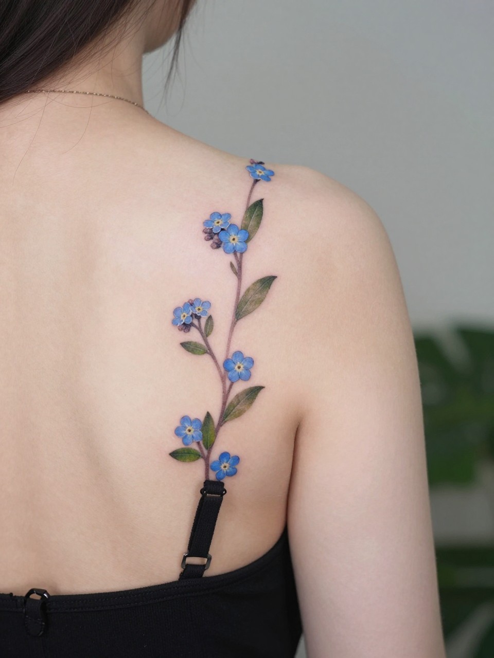 Angled back shot tattoo: small clusters of forget-me-nots and leaves scattered along the line where a bra strap would sit. Delicate and intentionally placed for reveal."
