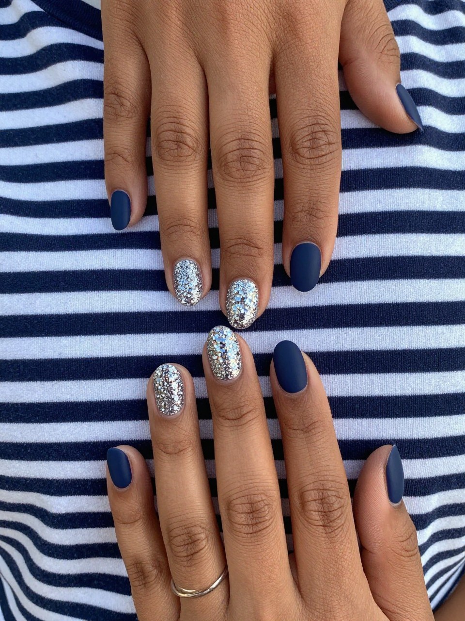 Top-down view of black feminine hands with short, oval nails resting on a navy blue and white striped shirt. Most nails are a matte navy blue. One ring finger is covered in dense, sparkling silver glitter with a glossy top coat. Daylight catches the sparkle beautifully.