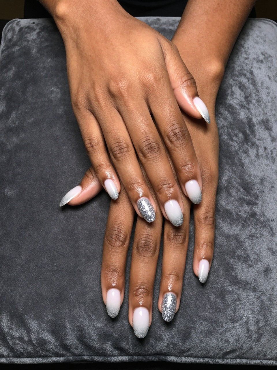 Bird's-eye view of black feminine hands laid flat on a dark, slate grey velvet cushion. The oval nails feature a smooth ombré from pure white at the cuticles to a dense concentration of sparkling silver glitter at the tips. Spotlight lighting makes the glitter tips dazzle dramatically.