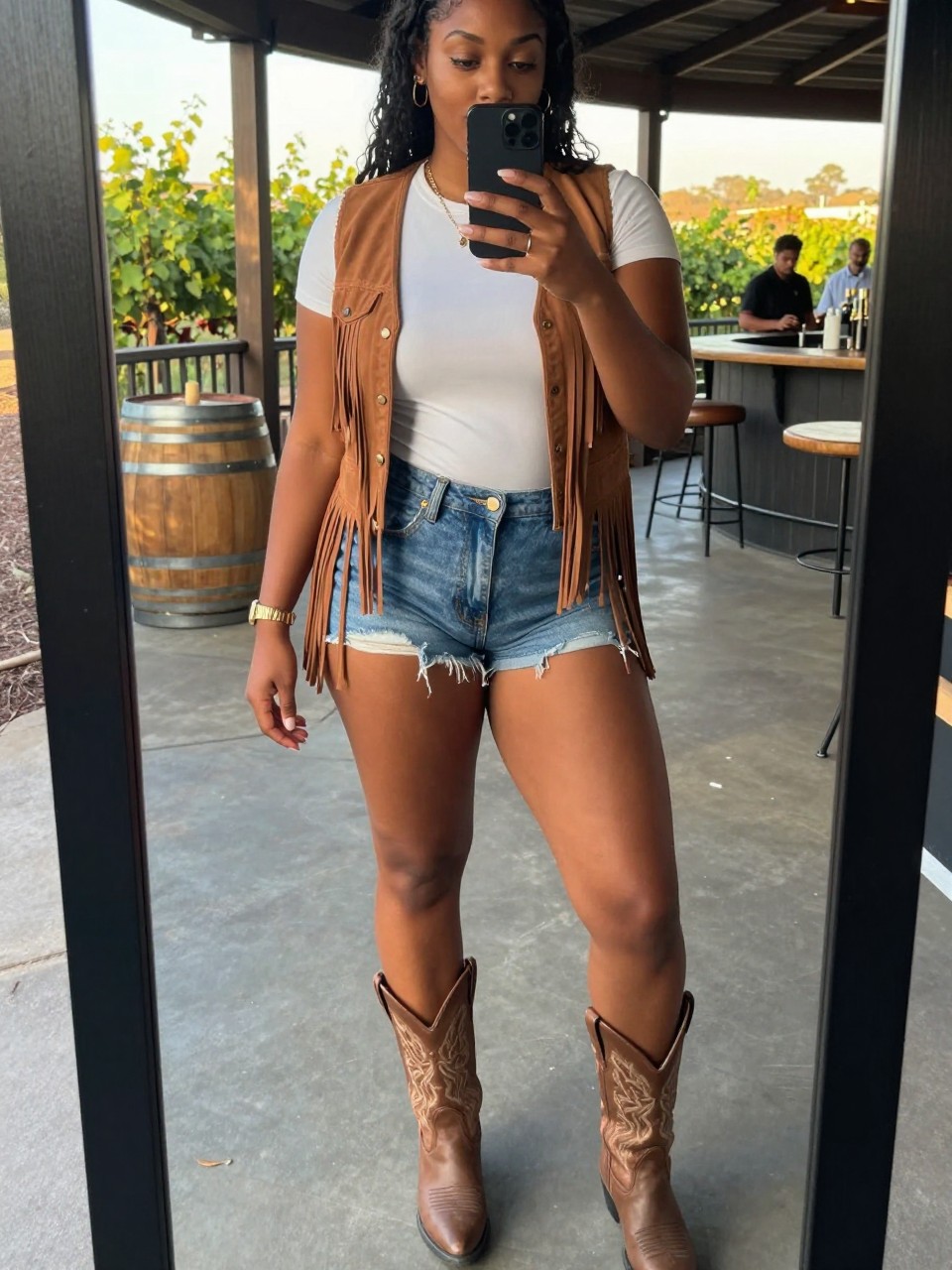 mirror selfie of a curvy black woman in a fringe vest over a white tee with denim shorts and cowboy boots, standing at an outdoor venue or bar, cropped at NECK so face doesn't show, at a winery, evening golden hour, iPhone photo quality.