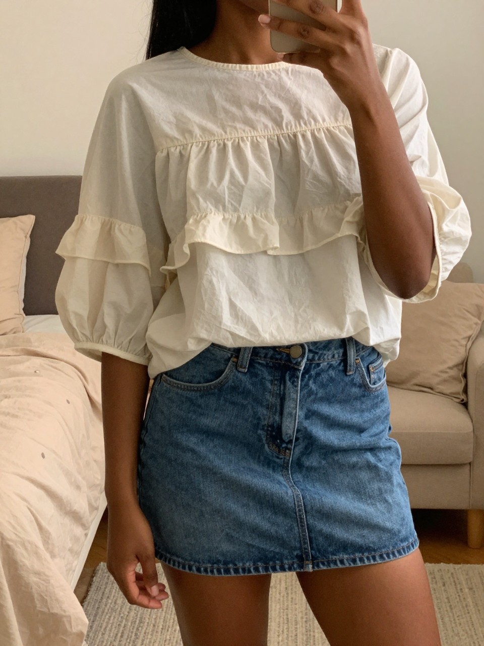 mirror selfie of a black woman in a tiered-sleeve blouse and denim skirt, showing off the sleeve detail, cropped at NECK so face doesn't show, in a feminine-themed bedroom, warm moody lighting, iPhone photo quality.