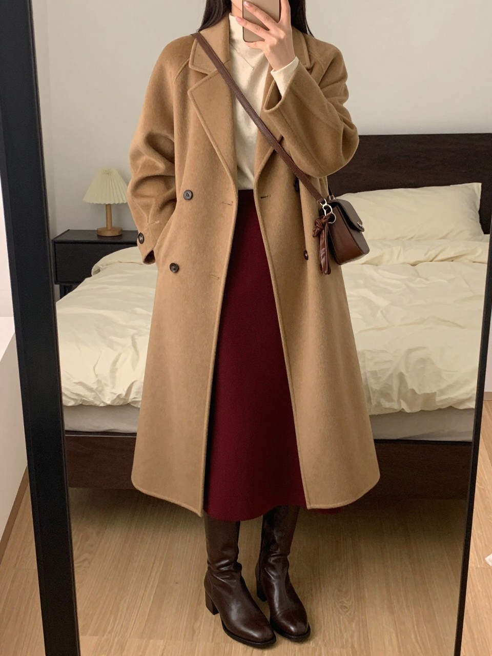 mirror selfie of a woman wearing a long camel wool coat over a cream top tucked into a burgundy red midi skirt with brown leather knee-high boots and a matching brown crossbody bag, holding leather gloves, standing in an elegant winter pose, cropped at NECK so face doesn't show, in a feminine-themed bedroom, warm moody lighting, iPhone photo quality.