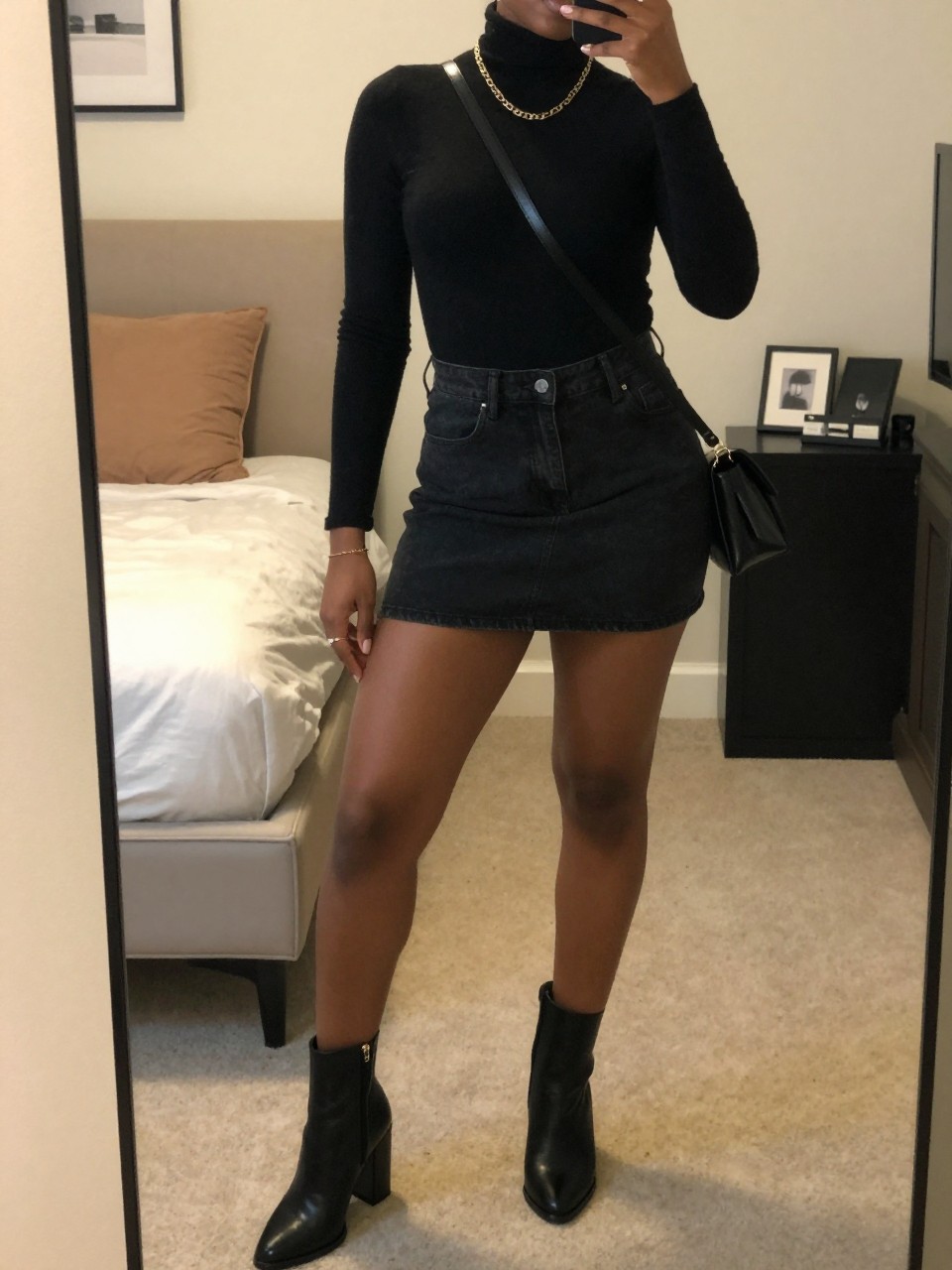 mirror selfie of a black woman wearing a black turtleneck tucked into a black denim mini skirt with black block-heel ankle boots and a gold chain necklace, holding a black crossbody bag, standing in an elegant pose, cropped at NECK so face doesn't show, in a feminine-themed bedroom, warm moody lighting, iPhone photo quality.