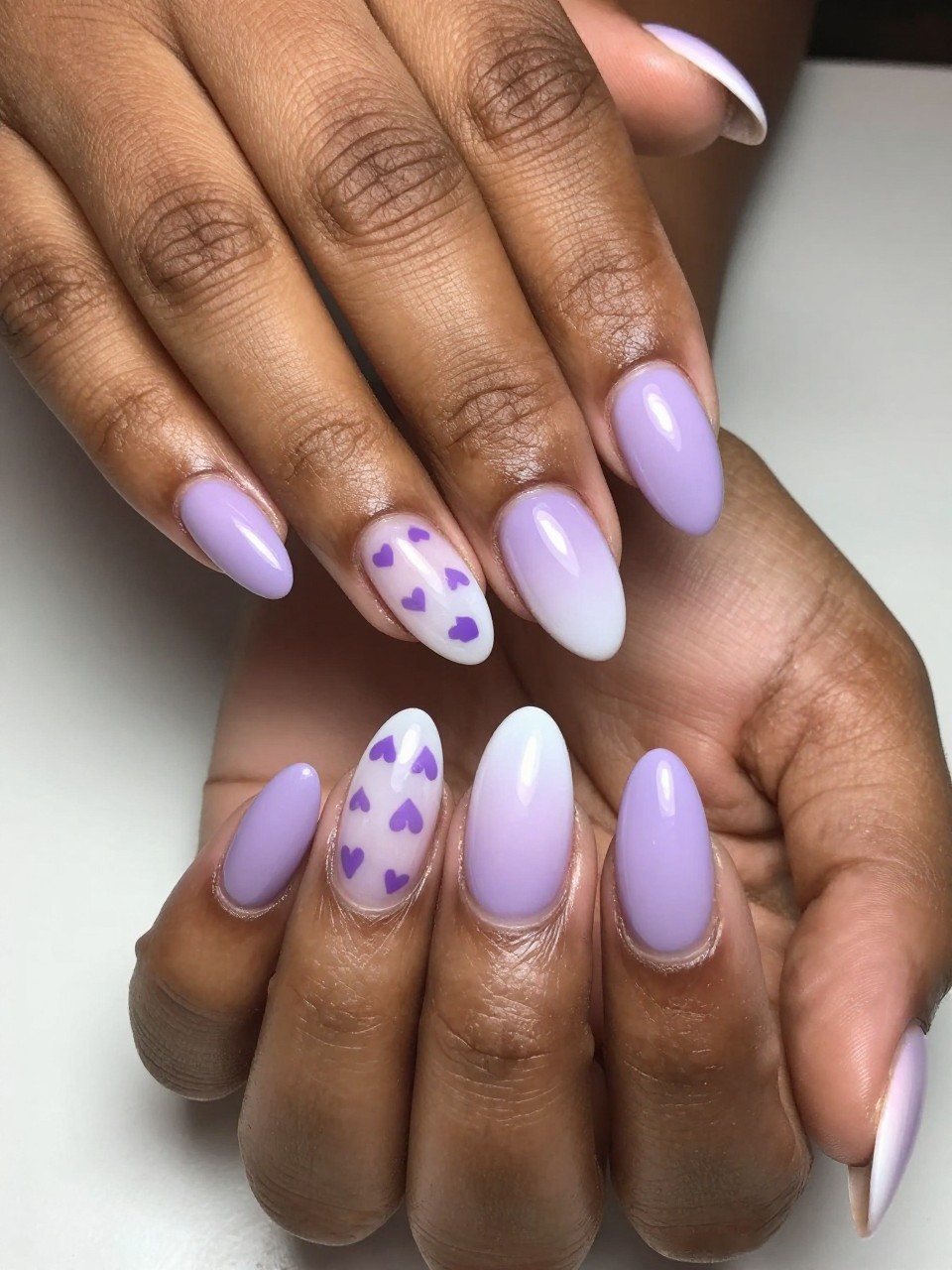 Angled view of black feminine hands with almond nails. The nails show a soft lilac-to-white ombré. Small purple hearts are painted along the gradient, becoming more transparent near the tip. The effect is soft, gradient, and beautifully seamless.