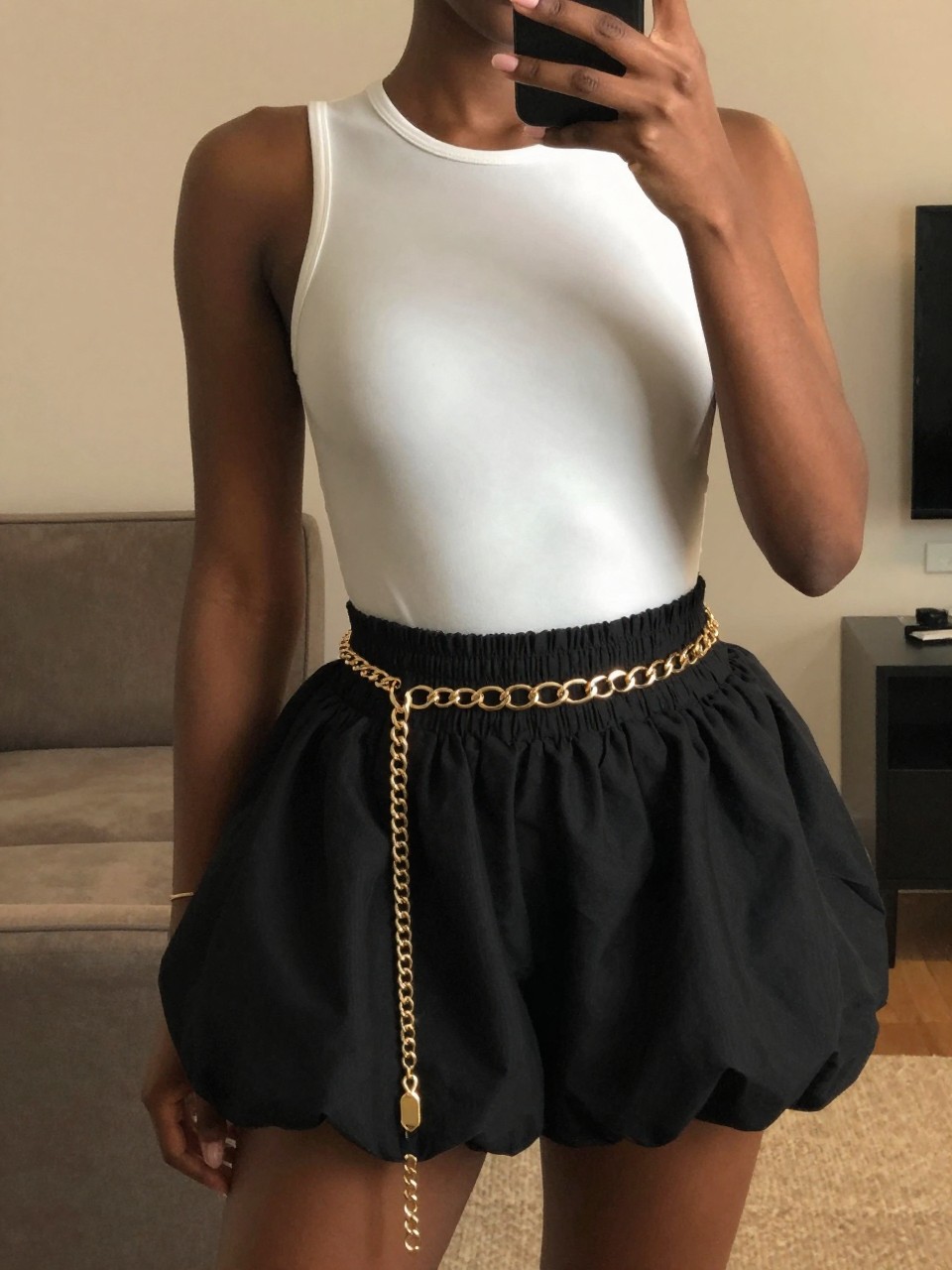 iPhone mirror selfie of a black woman in a white bodysuit and a black bubble skirt with a gold chain belt, hand resting on the chain, cropped at NECK so face doesn't show, in a city apartment, warm moody lighting, iPhone photo quality.