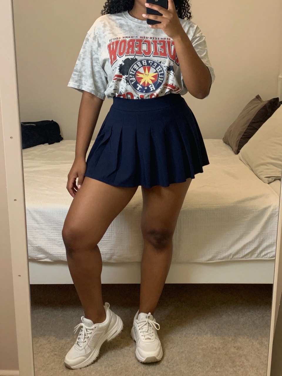 mirror selfie of a curvy black woman wearing an oversized vintage band tee with a navy tennis skirt and chunky white sneakers, holding her phone low by her thigh, cropped at NECK so face doesn't show, in a feminine-themed bedroom, warm moody lighting, iPhone photo quality.