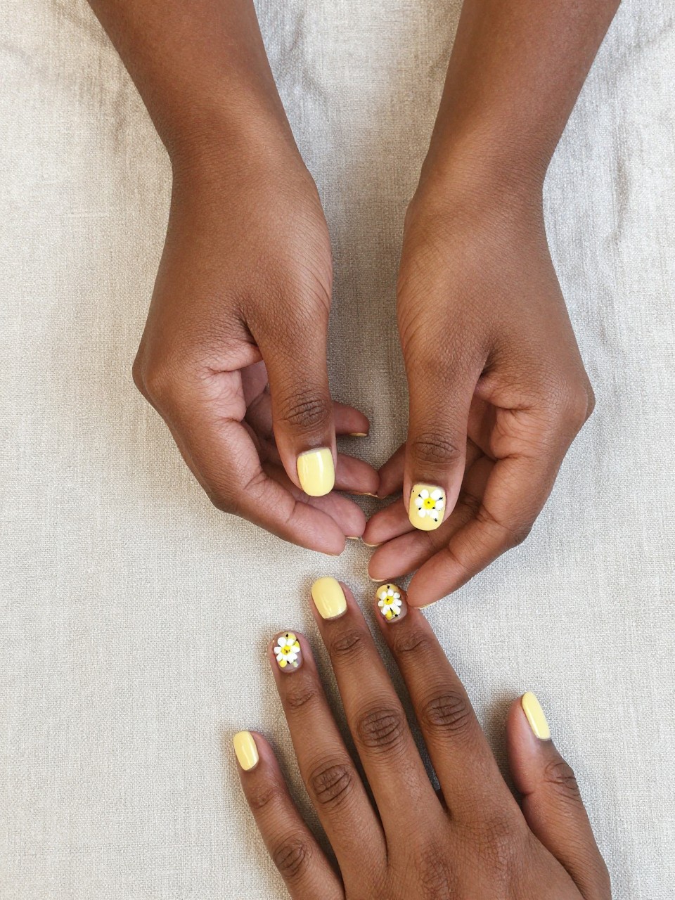 Overhead view of black feminine hands with short nails resting on a light, natural linen cloth. The nails are a pale chamomile yellow. Small white and yellow flowers are painted with tiny black “stitch” marks. The vibe is cozy, handmade, and sweet.