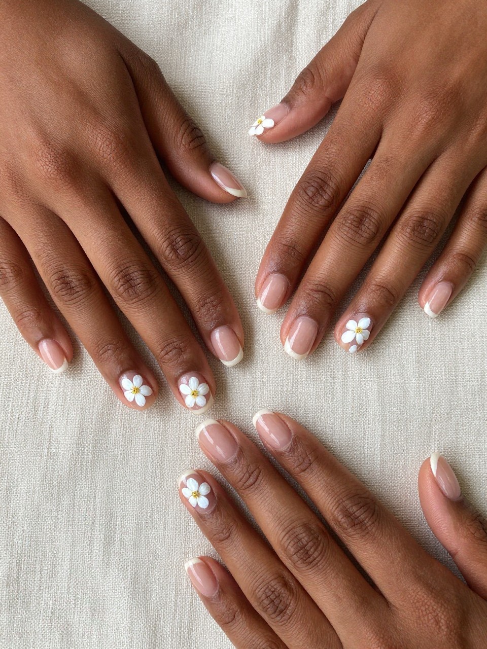 Overhead, top-down view of black feminine hands with well-manicured, short nails resting on a beige linen cloth. The nails have a clear, glossy base and there are three or four micro white blossoms with pale yellow centers are scattered on each nail design. The lighting is soft and natural, emphasizing the delicate art.